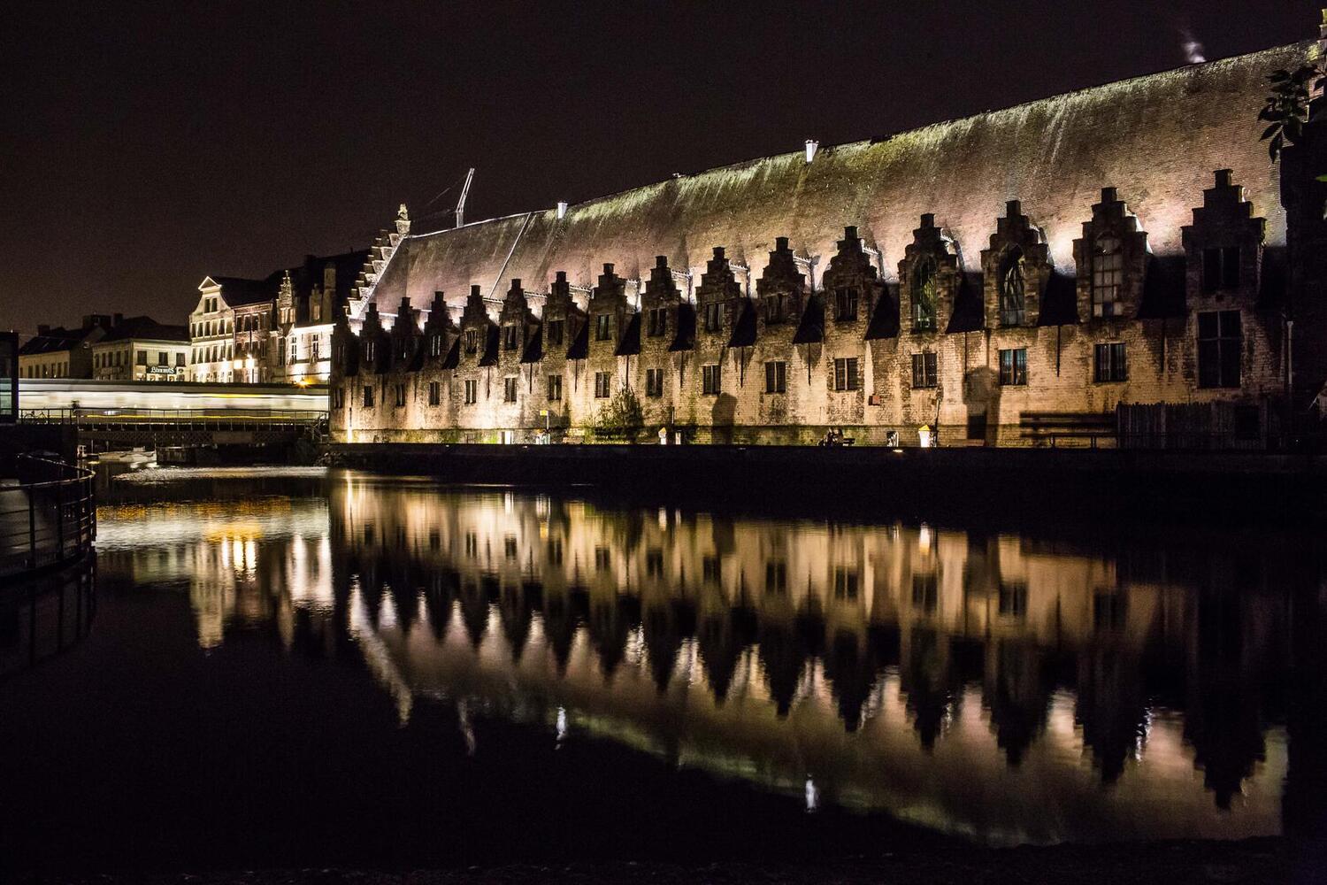 The medieval Great Butchers' Hall in Ghent at dusk, lit by warm interior light through stone window arches.