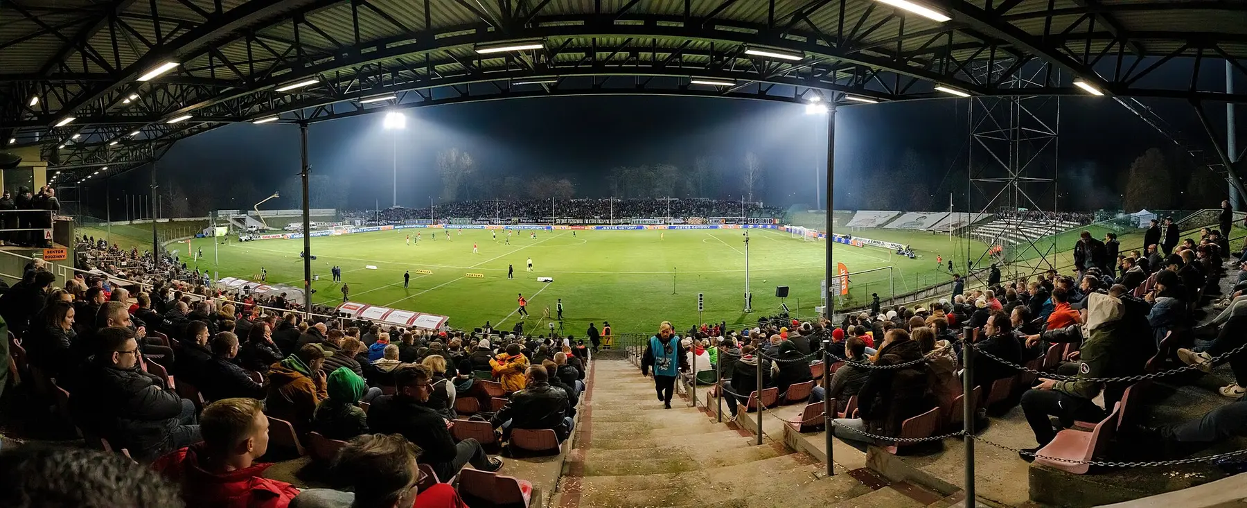 A floodlit football stadium at night, full stands, seen from behind the terrace steps